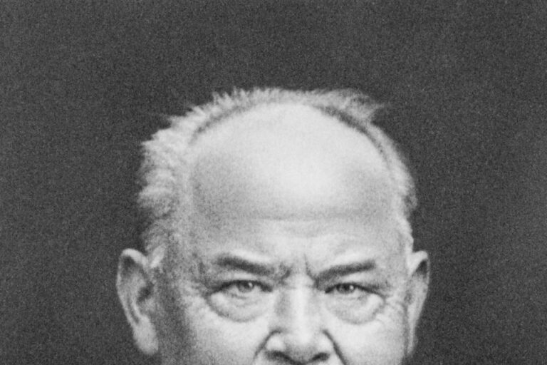 Schwarz-weiß-Porträt eines älteren Mannes mit Halbglatze, Schnurrbart, Anzug und Krawatte, ernster Blick in die Kamera.