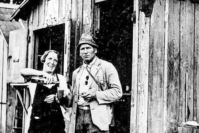 Eine Frau mit Schürze und ein Mann mit Mütze stehen vor einer Holzhütte im Wald.