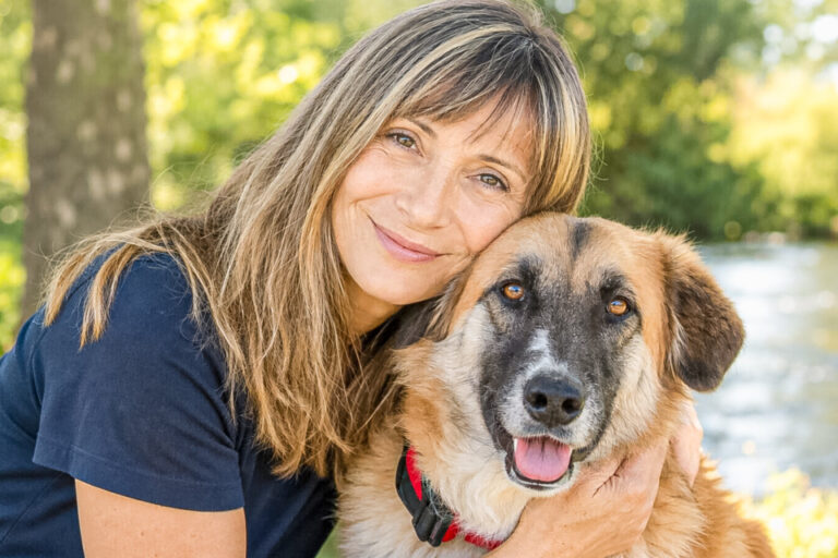 Eine lächelnde Frau umarmt liebevoll einen grossen braunen Hund im Freien an einem Fluss.