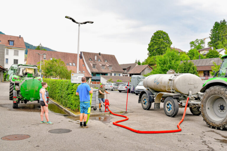 Personen befüllen einen Tankwagen mit rotem Schlauch neben grünen Traktoren auf einem Dorfplatz.