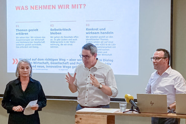 Drei Personen stehen an einem Rednerpult vor einer Leinwand mit Präsentationsfolie im Vortragssaal.