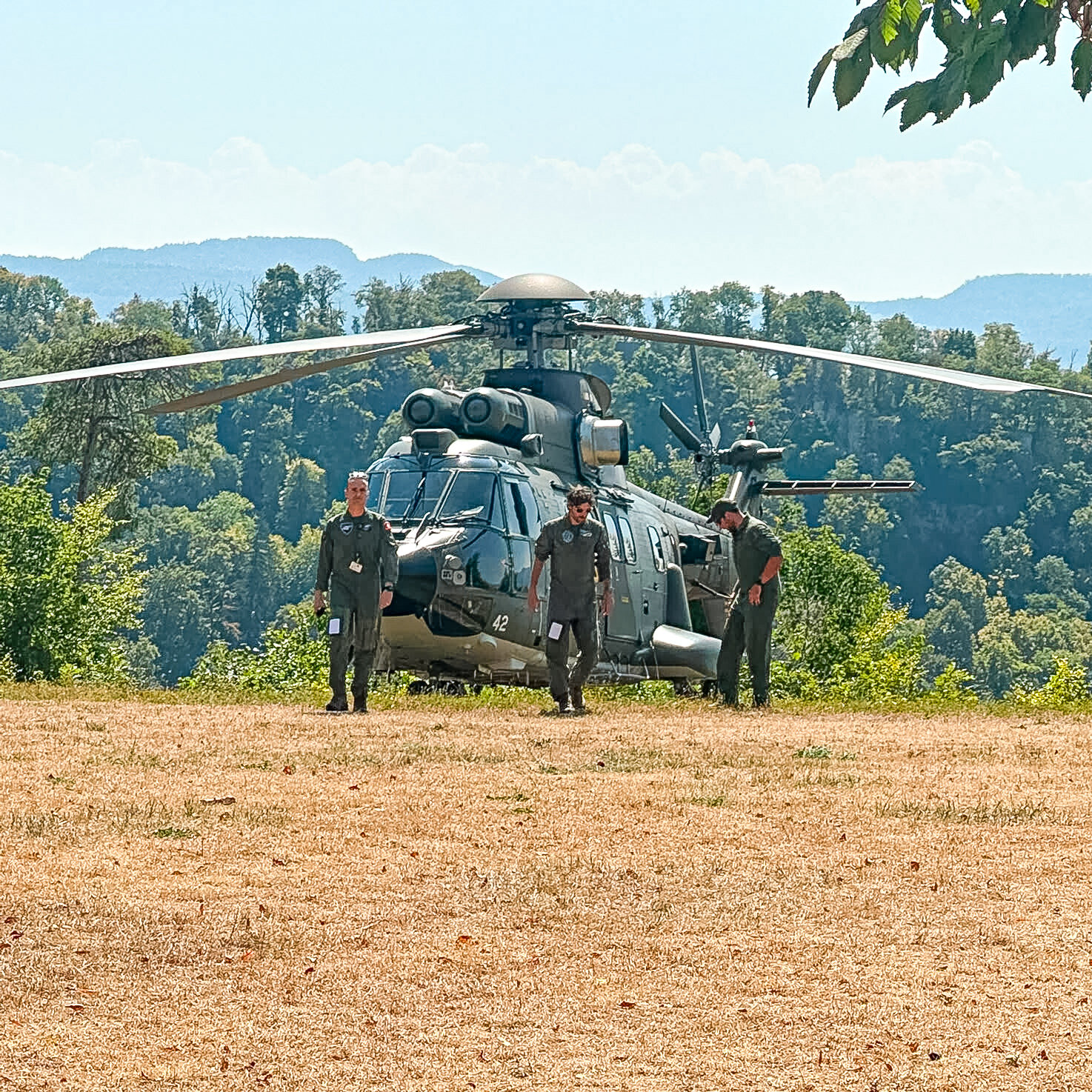 Drei Soldaten stehen vor einem grossen Militärhelikopter auf einer trockenen Wiese vor bewaldeten Hügeln.