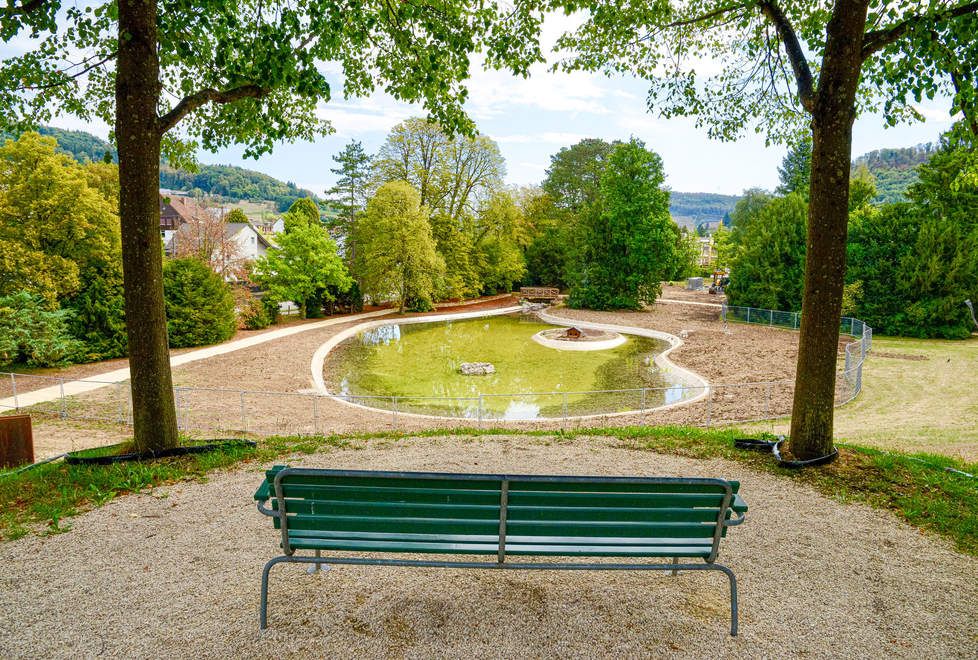 Grüne Parkbank vor einem kleinen Teich in einer Grünanlage mit Bäumen und Häusern im Hintergrund.