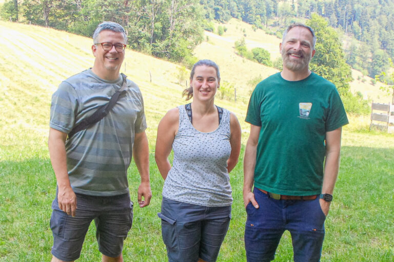 Drei lächelnde Erwachsene stehen nebeneinander auf einer grünen Wiese vor bewaldeten Hügeln.