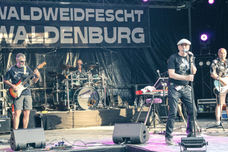 Eine Band spielt live auf einer Bühne mit Gitarren, Schlagzeug und Gesang vor einem Bühnenbanner.