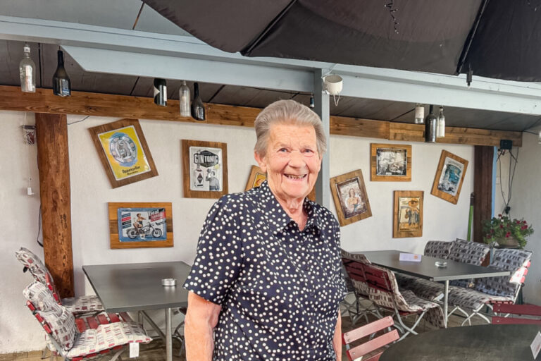 Eine ältere Frau mit gepunkteter Bluse lächelt in einer überdachten Restaurantterrasse.