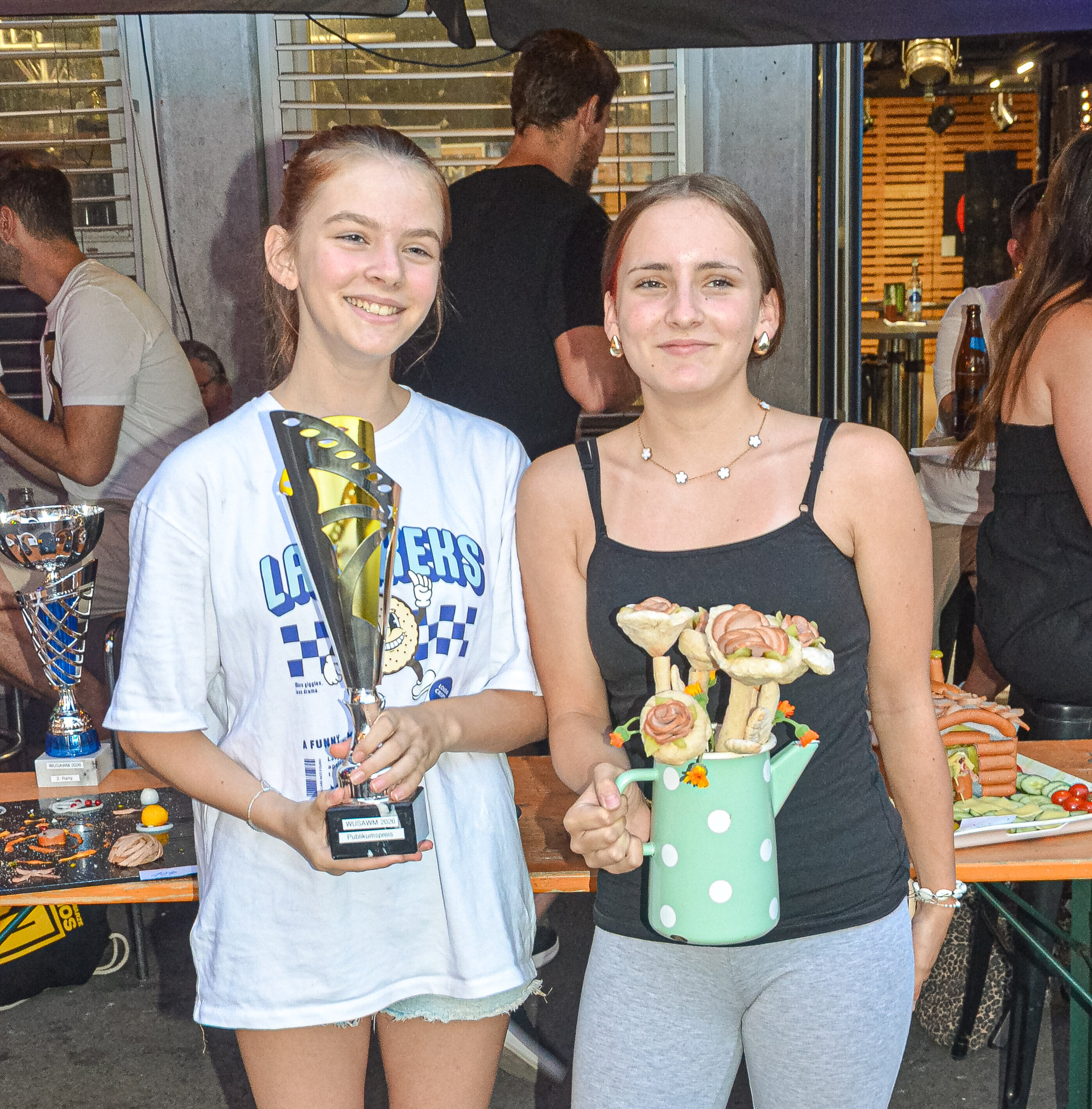 Zwei lächelnde junge Frauen stehen nebeneinander, eine hält einen Pokal, die andere ein Gefäß mit Spießen.