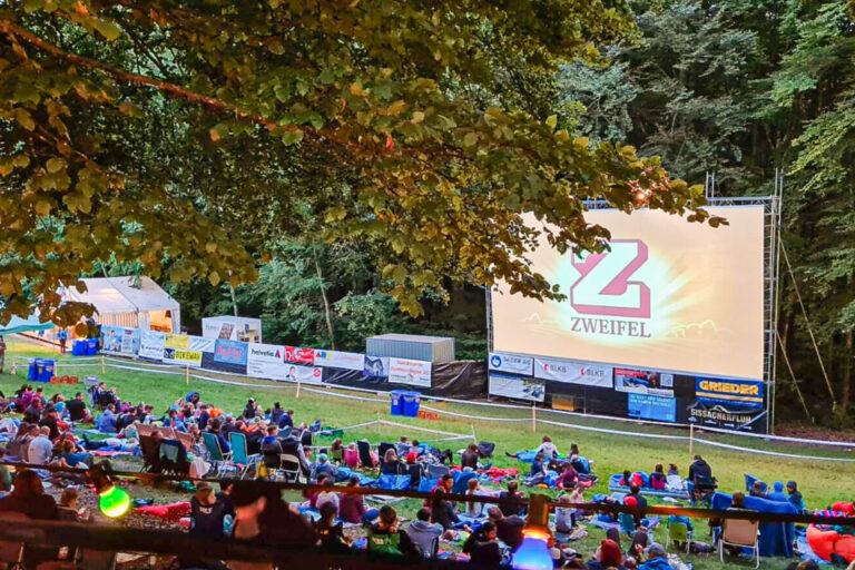 Zuschauer sitzen auf Decken im Freien vor einer Leinwand mit Werbebanner unter Bäumen bei Dämmerung.